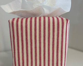 May include: A square fabric tissue box cover featuring a vertical red and cream-coloured ticking stripe pattern. The cover is constructed from a woven textile material and is shown with a white paper tissue extending from the top opening.