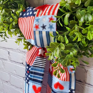 May include: A red, white, and blue fabric ribbon with stars and hearts, tied in a bow around a green wreath. The ribbon is made of patchwork fabric with different patterns.