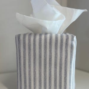 Könnte beinhalten: Rechteckige Taschentuchbox-Hülle mit Stoffbezug. Der Stoff hat einen weißen Hintergrund mit vertikalen grauen Streifen. Weiße Taschentücher ragen aus der Oberseite heraus. Ein schlichtes, elegantes Design.