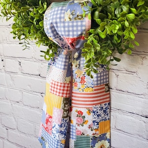 May include: A green wreath with a patchwork fabric bow. The bow features a mix of floral, gingham, and striped patterns in shades of blue, red, yellow, and white. The wreath is set against a white brick wall.