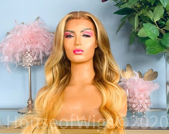 May include: A long, wavy, blonde wig with a light brown ombre effect. The wig is styled in loose waves and has a natural-looking parting. The wig is worn on a mannequin head.