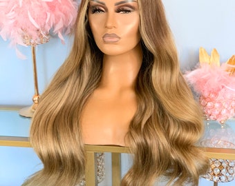 May include: A long, wavy, light brown wig with blonde highlights. The wig is styled with loose waves and falls to the shoulders. The wig is on a mannequin head.