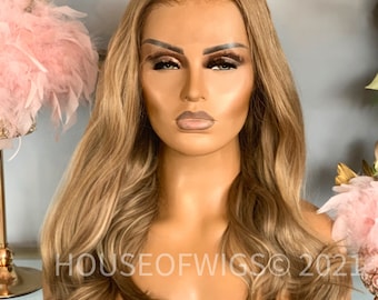 May include: A long, wavy, light brown wig with blonde highlights. The wig is styled with loose waves and falls to the shoulders. The wig is on a mannequin head.