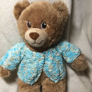 May include: A brown teddy bear wearing a blue and white knitted cardigan. The cardigan has a button closure and a speckled pattern.