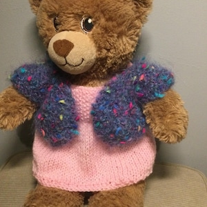 May include: A brown teddy bear wearing a pink knitted dress and a blue, fuzzy bolero jacket with colorful flecks. The bear has a brown nose and a smiling expression. The outfit is handmade and adds a touch of charm to the plush toy.