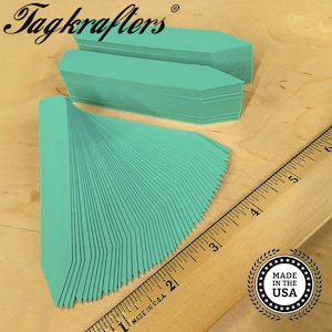 May include: A stack of teal-colored plant tags with a pointed end, fanned out on a wooden surface. The text "Tagkrafers" is in the upper left corner. A ruler and a "Made in the USA" emblem are also visible.
