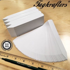May include: Stacks of white plant tags with a pointed end and a serrated edge. The image includes a ruler, a black pen, and the text "Tagkrafers" and "MADE IN THE USA". The tags are on a wooden surface.