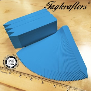 May include: A stack of 100 blue plastic plant markers with pointed ends. The markers are arranged in a fan shape and measure approximately 6 inches in length. The text "Tagkraftes®" is visible in the top right corner of the image. A ruler is visible in the bottom of the image.