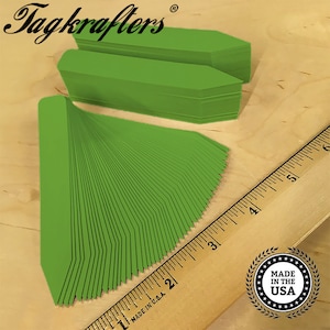 May include: A stack of 50 green plant markers, each measuring approximately 2.5 inches long, with a pointed tip. The markers are arranged in a fan shape, with the tips pointing outwards. A ruler is visible in the background, with the numbers 2, 3, 4, and 5 visible. A sticker with the text "MADE IN THE USA" is also visible in the background.