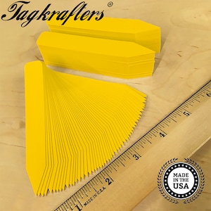 May include: A stack of 100 yellow plastic plant markers with pointed ends. The markers are approximately 2 inches long and are arranged in a fan shape. A ruler is in the background to show the size of the markers. The text "Tagkrafers" is visible in the top left corner of the image. The text "Made in the USA" is visible in the bottom right corner of the image.