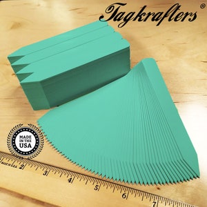 May include: Stacks of teal plant tags with a pointed top and a serrated bottom edge, arranged on a wooden surface. A ruler and a "Made in the USA" emblem are visible. The brand name "Tagkrafers" is displayed in elegant script.