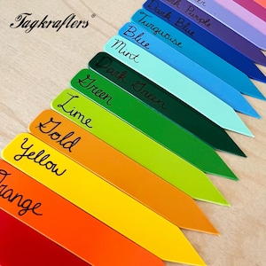 May include: A collection of colorful plant markers in various shades, including orange, yellow, green, and blue. Each marker has a rounded top and a pointed end, with handwritten labels indicating the color name. The Tagkrafers logo is visible in the upper left corner.