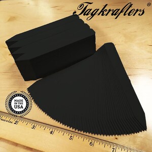 May include: Stacks of black plant markers with a pointed end, fanned out on a wooden surface. The text "Tagkrafers" is at the top. A ruler and a "Made in the USA" emblem are also visible.