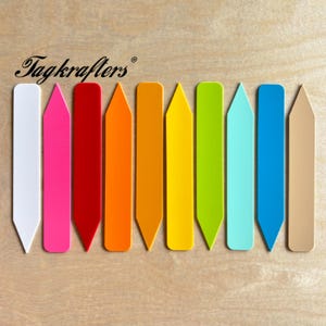 May include: A collection of colorful plant markers in various shades, including white, pink, red, orange, yellow, green, blue, and tan. The markers are arranged on a light wood surface, with the "Tagkrafters" logo in the upper left corner.