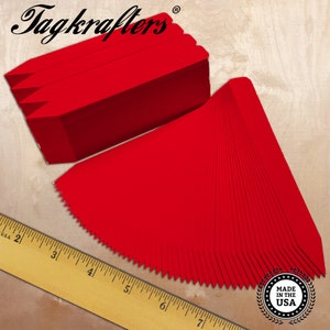 May include: Red paper tags fanned out and stacked, with a ruler in the foreground. The tags have a pointed end and are arranged on a light wood-grain surface. The text "Tagkrafters" is at the top left.