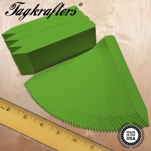 May include: A stack of bright green plant markers with a pointed top, fanned out on a wooden surface. The brand name "Tagkrafters" is in the upper left corner. A wooden ruler is in the lower left corner, and a "Made in the USA" seal is in the lower right.
