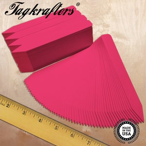 May include: A stack of bright pink plant tags with a pointed end and a scalloped edge. The brand name "Tagkrafters" is printed in black script at the top. A wooden ruler and a "Made in the USA" seal are also visible.
