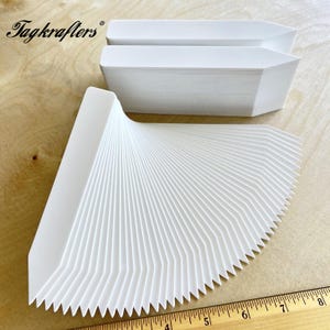 May include: White paper plant markers with a pointed top and a serrated bottom edge, fanned out on a wooden surface. The brand name "Tagkrafers" is visible in the upper left corner. A ruler is in the bottom right corner.