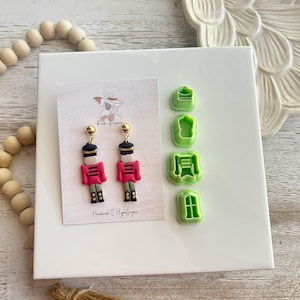 May include: A pair of handmade nutcracker earrings with gold accents and red and white details. The earrings are displayed on a white background with green plastic cookie cutters.