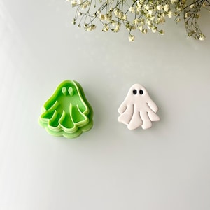 May include: A green plastic ghost cookie cutter and a white ceramic ghost cookie. The cookie cutter is open and the cookie is solid.