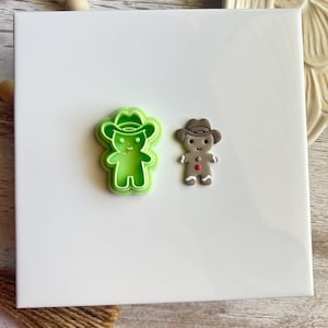 May include: Two cookie cutters, one green and one brown, shaped like gingerbread people wearing cowboy hats. The green cutter is a cutout design, while the brown cutter is solid.