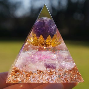 May include: A clear resin pyramid with a purple amethyst sphere and a gold-colored lotus flower. The pyramid is filled with amethyst fragments, glitter, and gold flakes. The pyramid is held in a hand.