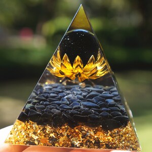 K&ouml;nnte beinhalten: Eine klare Pyramide mit einer goldenen Lotusblume und schwarzen Obsidian-Steinen im Inneren. Die Pyramide ist aus klarem Kristall gefertigt und hat eine goldene Basis.