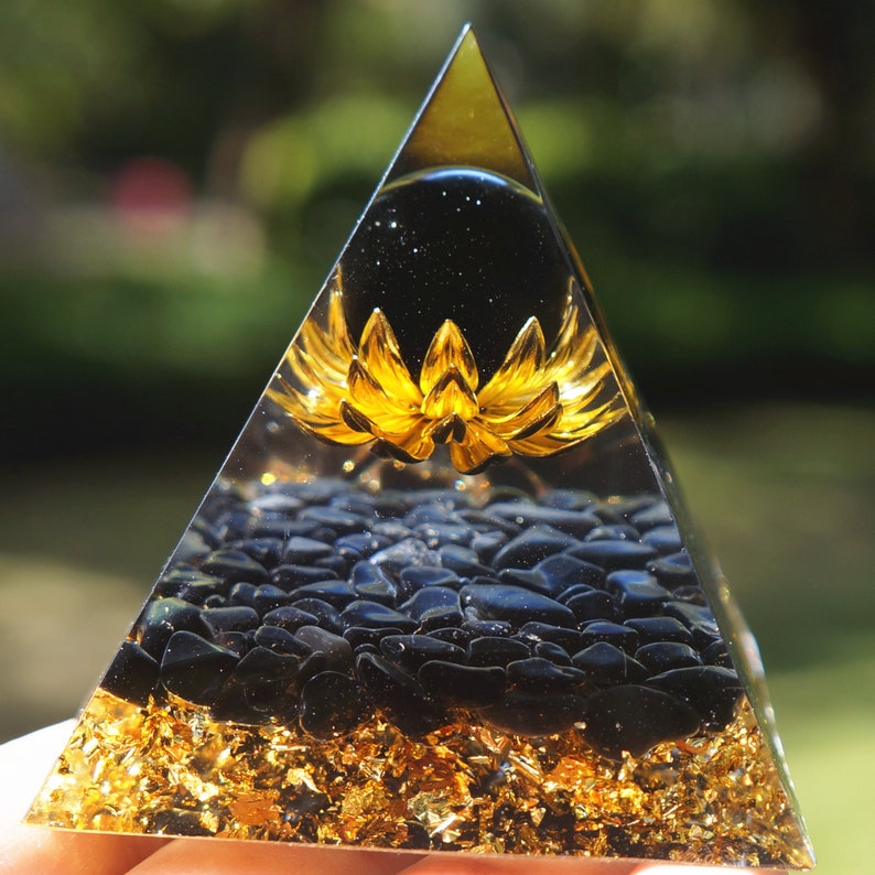 K&ouml;nnte beinhalten: Eine klare Pyramide mit einer goldenen Lotusblume und schwarzen Obsidian-Steinen im Inneren. Die Pyramide ist aus klarem Kristall gefertigt und hat eine goldene Basis.