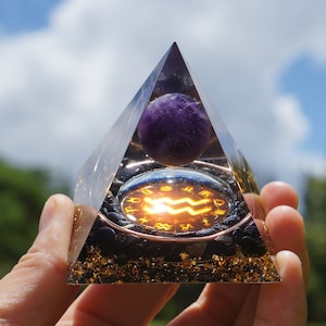 May include: A clear glass pyramid with a purple amethyst sphere inside. The base of the pyramid is filled with black and gold stones. The sphere is centered over a circular metal plate with the astrological symbol for Aquarius.
