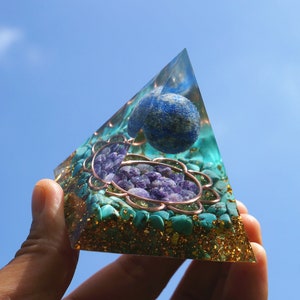 Puede incluir: Una pirámide de cristal transparente con una esfera azul, cristales de amatista púrpura y piedras turquesa en su interior. La pirámide se sostiene en una mano contra un cielo azul.