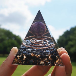 May include: A clear crystal pyramid with a copper wire spiral and black obsidian stones. A purple amethyst sphere sits at the top of the pyramid.