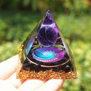 Pode incluir: Uma pirâmide transparente com uma base de glitter dourado, contendo uma esfera de ametista roxa e um design espiral azul e roxo.