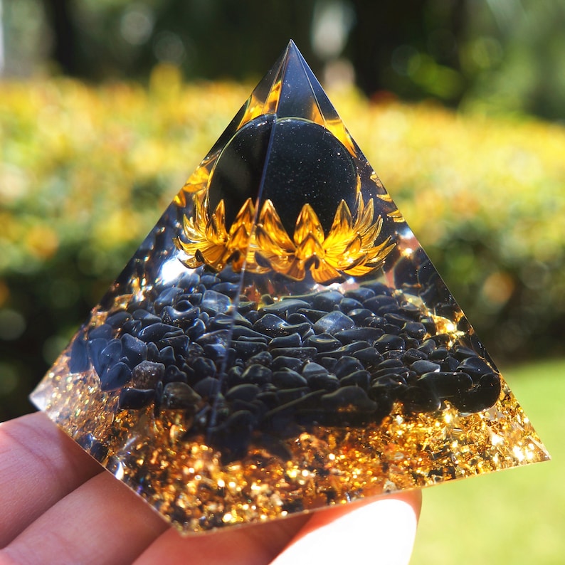 K&ouml;nnte beinhalten: Eine klare Glaspyramide mit einer schwarzen Obsidiankugel an der Spitze. Die Pyramide ist mit schwarzen Obsidiansplittern und Goldflocken gef&uuml;llt.