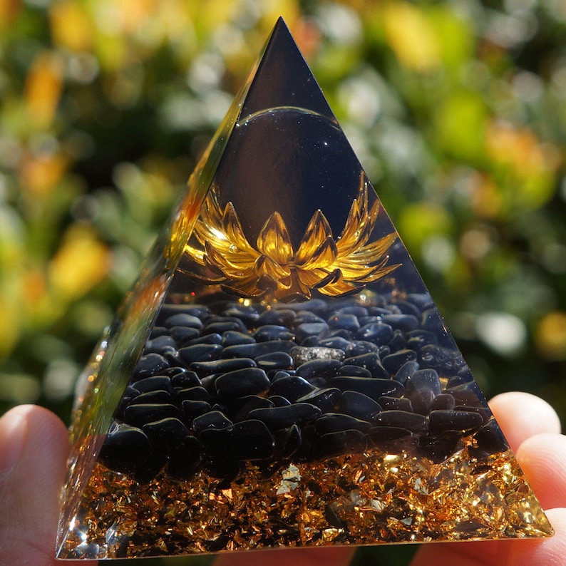 K&ouml;nnte beinhalten: Eine klare Glaspyramide mit einer goldenen Lotusblume im Inneren. Die Pyramide ist mit schwarzen Obsidian-Steinen und goldenen Flocken gef&uuml;llt.