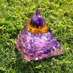 May include: A purple amethyst pyramid with a gold flower design and a purple amethyst sphere in the center. The pyramid is sitting on a bed of green grass.