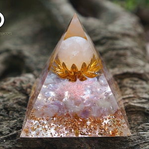 Könnte beinhalten: Eine transparente pyramidenförmige Orgon-Pyramide mit einer goldenen Lotusblume und einer rosafarbenen Kristallkugel im Inneren. Die Pyramide steht auf einer braunen Holzoberfläche.  "Infinite Orgone" ist auf der Oberfläche geschrieben.