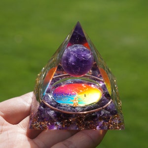 Puede incluir: Una pirámide transparente hecha de cristales de amatista con una esfera de amatista púrpura en su interior. La pirámide tiene un alambre de cobre envuelto alrededor y un diseño de mandala colorido en su interior.