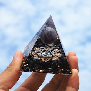 Puede incluir: Una pirámide de cristal transparente con un diseño de metal negro y dorado en su interior. La pirámide está llena de piedras negras y escamas doradas. Una esfera de amatista púrpura se asienta en la parte superior de la pirámide.
