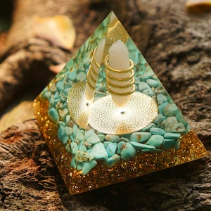 Peut inclure: Une pyramide en cristal transparente avec une spirale en métal doré et une pointe de cristal blanc à l'intérieur. La pyramide est remplie de petites pierres turquoise et de paillettes dorées.