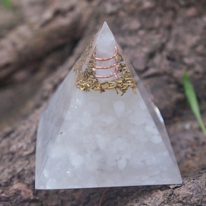 May include: A clear quartz pyramid with embedded copper coils and gold-coloured metal accents. The pyramid is set against a blurred natural background, showcasing its geometric form and decorative elements. The pyramid is a decorative item.