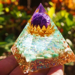 May include: A clear pyramid-shaped crystal with a purple amethyst crystal at the top, surrounded by gold-coloured metal. The pyramid is filled with small turquoise stones and gold glitter.