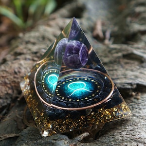 May include: A clear crystal pyramid with a purple amethyst sphere at the top. Inside the pyramid is a copper wire wrapped around black stones and a teal blue symbol with gold stars.