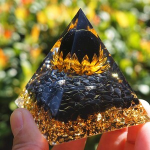 K&ouml;nnte beinhalten: Eine schwarze Obsidian-Kristallpyramide mit goldenen Blattverzierungen und einer schwarzen Obsidiankugel an der Spitze. Die Pyramide ist mit schwarzen Obsidiansplittern gef&uuml;llt und hat ein goldenes Blumendesign um die Kugel.