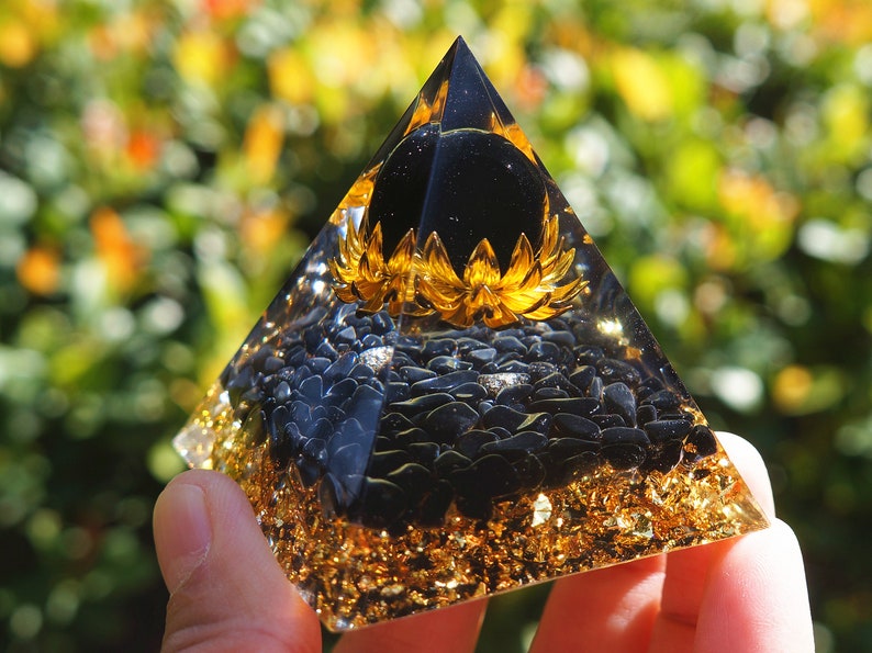 K&ouml;nnte beinhalten: Eine schwarze Obsidian-Kristallpyramide mit goldenen Blattverzierungen und einer schwarzen Obsidiankugel an der Spitze. Die Pyramide ist mit schwarzen Obsidiansplittern gef&uuml;llt und hat ein goldenes Blumendesign um die Kugel.