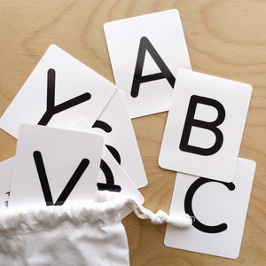 May include: White flashcards with black uppercase letters, including A, B, C, and V, are scattered on a light wood surface. A small white drawstring bag holds additional cards. The cards are a learning tool for children.