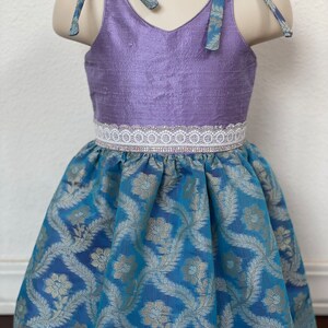 May include: A purple sleeveless dress with a white lace trim and a blue floral print skirt. The dress has two blue fabric ties that can be tied at the shoulders.