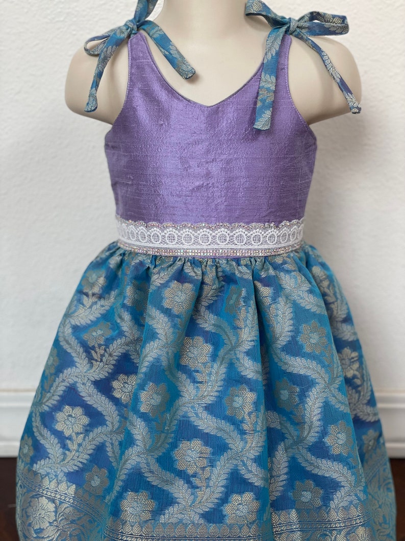 May include: A purple sleeveless dress with a white lace trim and a blue floral print skirt. The dress has two blue fabric ties that can be tied at the shoulders.
