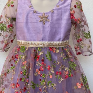 May include: A lavender dress with a floral embroidered tulle skirt and a satin bodice. The bodice has a star-shaped embellishment and a gold trim around the waist.