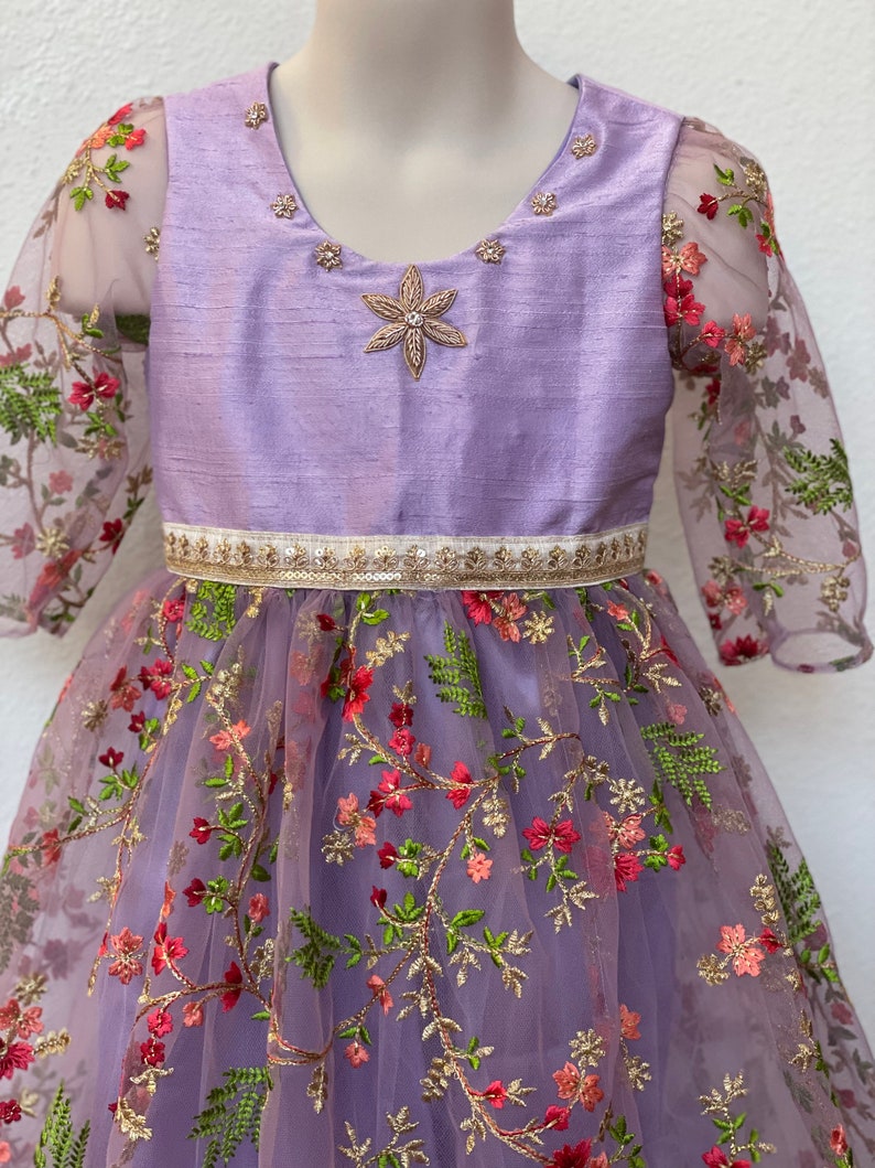 May include: A lavender dress with a floral embroidered tulle skirt and a satin bodice. The bodice has a star-shaped embellishment and a gold trim around the waist.