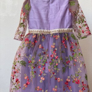 May include: A lavender dress with a floral embroidered tulle overlay. The dress has a round neckline and 3/4 sleeves. The bodice is made of a solid lavender fabric and the skirt is made of tulle with embroidered flowers in shades of pink, green, and gold.